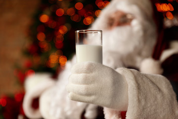 Santa Claus holding glass of milk against blurred Christmas lights, closeup
