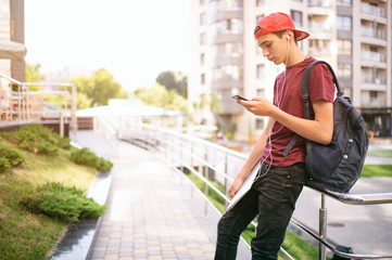 Fototapeta na wymiar Young man stands with backpack and holds smartphone, in the city. Teenage boy is using mobile phone, outdoors. Caucasian teenager in casual clothes with cell phone, urban scene.