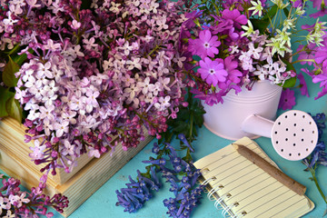 Lilac and spring flowers with diary book and watering can on working table.