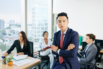 Serious confident Asian entrepreneur standing with arms folded, his employees working in background