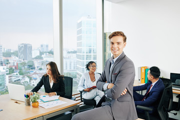 Handsome young smiling businessman sitting on desk with arms folded in modern office with big panoramic windows