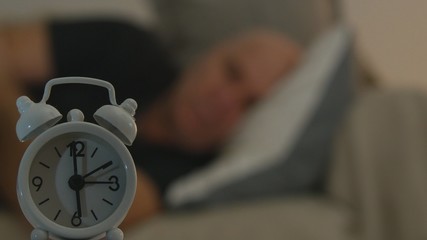 Tired Man Sleeping in the Bad with a Clock Alarm on the Table