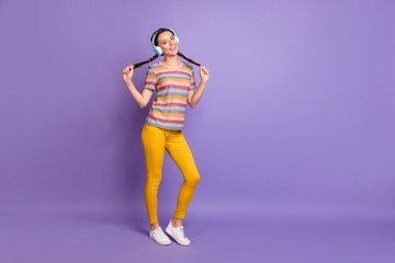 Fototapeta na wymiar Full length photo of positive cheerful girl relax rest listen rhythmical melody music from blue wireless headset enjoy hold braids wear stylish clothes isolated violet color background