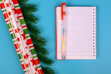 Christmas background. An open notebook for recording expenses. A pen. Packing colored paper. Fir branches. Blue background. Top view. Close-up. Place for text.