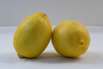 Two delicious lemons on white table cloth with gray background. Organic lemon leaning on other lemon. Fresh citrus fruit.