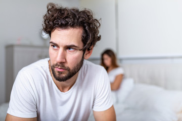 Sad thoughtful man after arguing with girlfriend.Relationship difficulties, conflict and family concept,unhappy couple having problems at bedroom. Sad guy sitting on bed,girlfriend in the background - obrazy, fototapety, plakaty