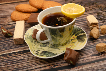 A Cup of black tea, tea leaves, pieces of brown sugar, oatmeal cookies, waffles, pieces of chocolate on a wooden background. Close up.