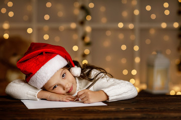 Happy little child girl in santa hat writing wish list or letter to Santa Claus on background with lights. Merry Christmas