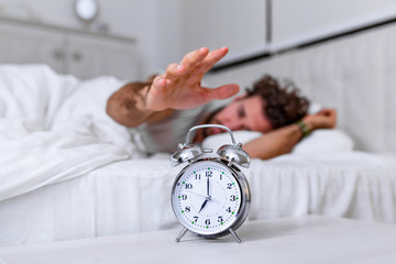 Man lying in bed turning off an alarm clock in the morning at 7am. Hand turns off the alarm clock waking up at morning, man turns off the alarm clock waking up in the morning from a call. - obrazy, fototapety, plakaty