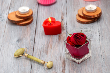 Massage, jade roller for the face, a burning candle in the shape of a heart, a rose in a glass cup on a gray wooden table. Valentine's Day. Personal care. Relaxation, aromatherapy at home.