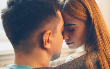 Close up side view portrait of a beautiful woman with red hair and freckles and her man sitting face to face with eyes closed before kissing at home.