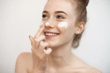 Foto op Canvas Portrait of a charming young female with red hair and freckles isolated on white applying a anti age cream on her face and nose smiling. © Strelciuc