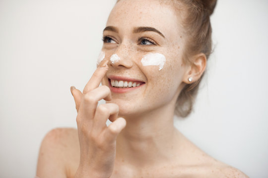 Portrait of a charming young female with red hair and freckles isolated on white applying a anti age cream on her face and nose smiling.