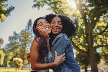 Smiling happy portrait of a diverse young black and white female friends hugging each other and having fun outdoors in the park  - obrazy, fototapety, plakaty