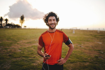 Portrait of a happy fitness young man in earphones holding mobile phone while standing in park...