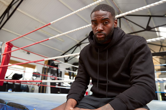 Portrait Of Male Boxer Wearing Hooded Sweatshirt In Gym Sitting On Boxing Ring - Powered by Adobe