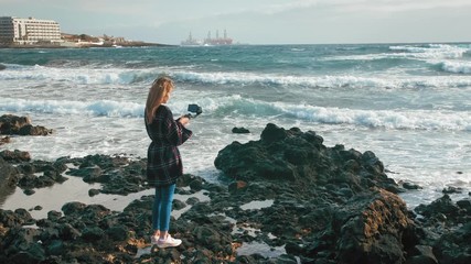 female blogger, journalist, influencer with gimbal make a report on pollution environment. Shoots video on a smartphone. oil rigs in the sea offshore on background. Lava stones. - Powered by Adobe