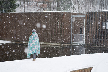 Girl in grey hat, covered in blue patterns plaid enjoying snowfall standing backfront near river dam