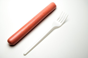 Sausage next to a fork on a white background. Copy space. The concept of fast food, GMOs, hunger or lack of food. Environmental issue and the ban on plastic dishes.