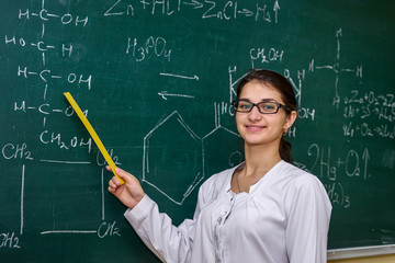 Woman standing in chemist's classroom near board and pointing on it
