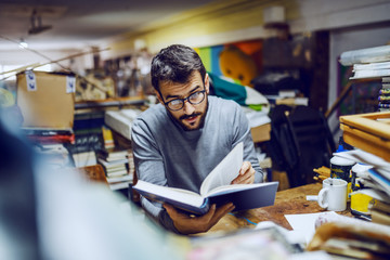 Young attractive caucasian bearded serious man with eyeglasses sitting in back of bookstore and...