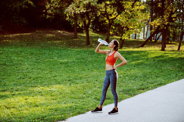 Attractive thirsty caucasian fit brunette in sportswear and with ponytail standing in nature and drinking water. Sunny day in nature.