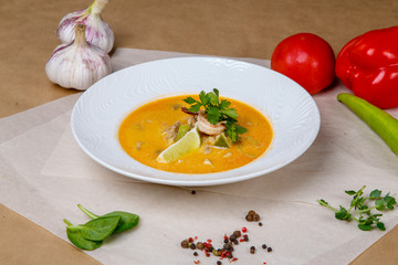 Cheese soup with shrimp. Yellow soup is decorated with lemon and parsley. White round plate lies on a white and gray wrapping paper, overlaid with pepper, peas, tomatoes, garlic, herbs. 