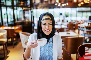 Charming modern smiling muslim woman sitting in cafe and holding cup of coffee. Next to her on chair are shopping bags. She is taking a break from all day shopping.