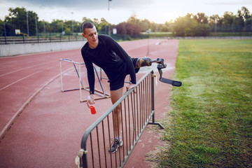 Full length of handsome fit caucasian sporty handicapped man in sportswear and with artificial leg leaping over the fence while holding refreshment in hand.