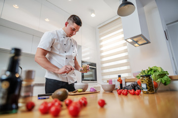 Low angle view of focused caucasian chef in uniform standing in kitchen and adding mustard in bowl. On kitchen counter are different sorts of vegetables.