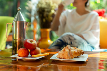 Blurred woman enjoys morning coffee with croissants and apple on wooden table.