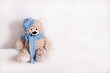 Children's toy teddy bear is sitting on a white sofa in a knitted scarf and hat in blue.