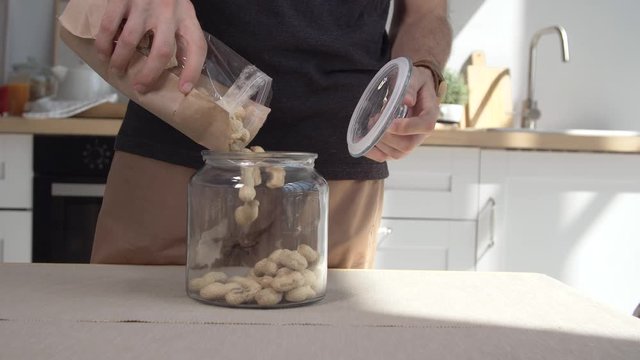 Man opens a glass jar and pours nuts from a paper bag into it. Young man shifts nuts in a glass jar at home kitchen. Healthy eating, vegetarian food, diet concept 