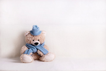 Children's toy teddy bear is sitting on a white sofa in a knitted scarf and hat in blue.