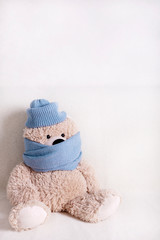 Children's toy teddy bear is sitting on a white sofa in a knitted scarf and hat in blue.