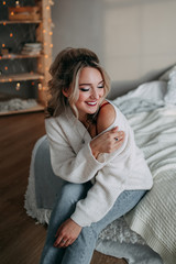 Fototapeta premium Attractive sexy happy blonde girl with a beautiful smile in a white jacket and jeans resting and relaxing on the bed in a cozy bright bedroom at sunset.