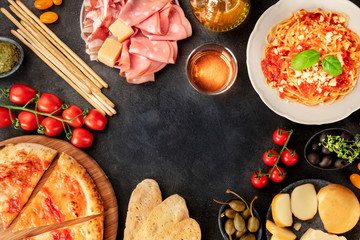 Italian food. Pizza, pasta, cheese, ham, wine, olives, pesto, olive oil, capers, shot from the top forming a frame with a place for text, a flat lay design template on a dark background