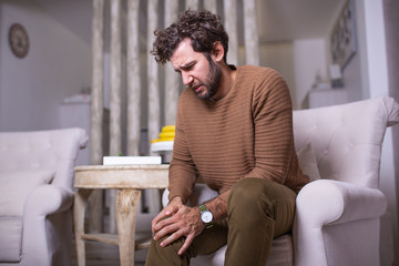 Young man suffering from knee pain sitting sofa at home. Man holding his leg and suffering from...