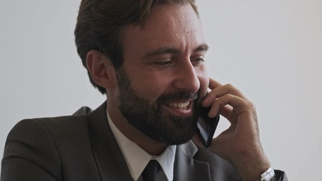 Close up view of happy handsome bearded businessman talking by smartphone while sitting at office