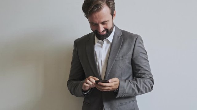 Happy handsome bearded businessman using smartphone while standing near the wall at office