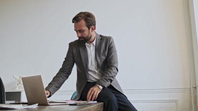 Serious handsome bearded businessman using laptop computer and writing something while sitting on the table at office