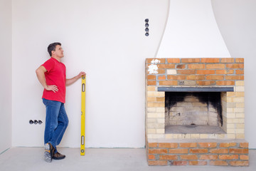 A white man stands with a yellow water level in his hands and looks thoughtfully at the unfinished fireplace.
