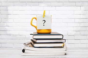 Yellow cup with a sticker question mark on a stack of notebooks and folders on the desktop on a brick wall background