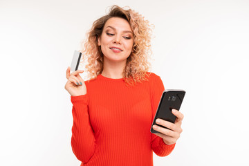 Naklejka premium curly blond surprised girl with a credit card in her hands looks at the phone against a white studio background