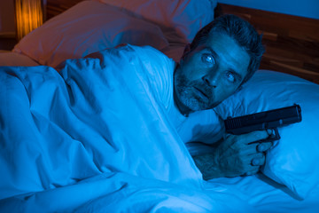 night edgy portrait of young stressed and paranoid American man lying on bed unable to sleep holding gun looking around scared suffering paranoia expecting intruders