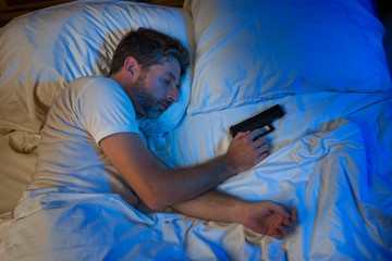 night portrait of young stressed and paranoid American man in bed sleeping and holding gun fearing...