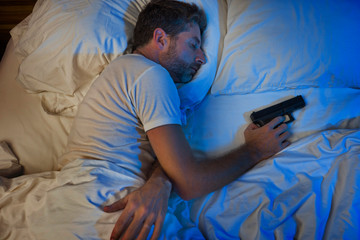 night portrait of young stressed and paranoid American man in bed sleeping and holding gun fearing...
