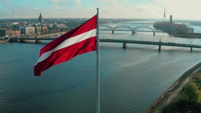 Latvian flag attached to a pole waving in the wind in front of the Daugava river and the skyline of Riga, Latvia