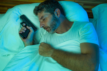 night portrait of young stressed and paranoid American man in bed sleeping and holding gun fearing...