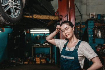 Fototapeta na wymiar The concept of small business, feminism and women's equality. A young woman mechanic wipes sweat from her forehead, on the lift is a car